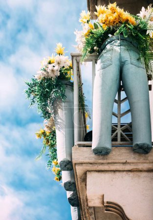Artistically arranged denim-covered legs protrude from an upper-level balcony, draped with vibrant yellow and white floral arrangements in a unique urban installation under a serene blue sky