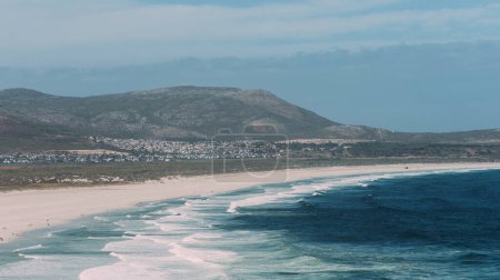 The tranquil scene captures the untouched beauty of Noordhoek Beach as the gentle waves wash over the pristine white sands. The backdrop of the rolling hills complements the peaceful beach, highlighting the natural splendor of South Africas coastline