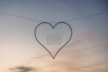 A delicate heart-shaped silhouette dangles from a thin wire, suspended against a soft gradient of twilight hues transitioning from day to night. The simplicity of the heart contrasts with the complexity of the skys natural palette.