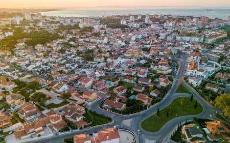 Photo for An aerial perspective captures the warm glow of sunset over the picturesque coastal town of Cascais, Portugal. The neatly laid-out residential areas with individual homes and gardens are intersected by curving roads, hinting at the peaceful, suburban - Royalty Free Image