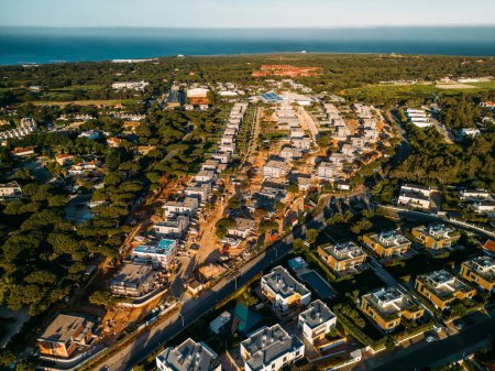 An aerial perspective showcases the luxurious residential district of Quinta da Marinha in Cascais, Portugal, bathed in the warm hues of the setting sun. The neatly lined homes and winding streets are surrounded by lush greenery, with the calm Atlant