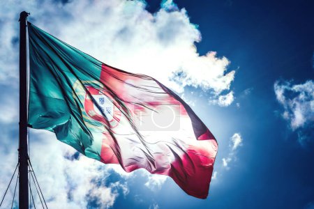 The vibrant colors of the Portuguese flag ripple in the breeze, proudly displayed against a striking backdrop of a clear blue sky. Sunlight filters through the fabric, creating a beautiful flare effect and highlighting the emblem at the flags center.