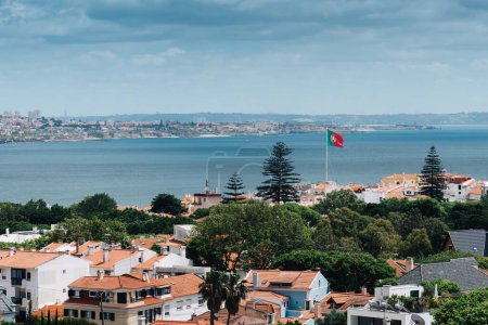 Un panorama panoramique surplombe la ville côtière de Cascais au Portugal, mettant en valeur des bâtiments blancs traditionnels avec des toits orange, dans un contexte océanique brumeux, avec le drapeau portugais lumineux