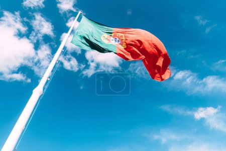 Das leuchtende Rot und Grün der portugiesischen Nationalflagge fällt auf, wenn sie im Wind auf einem hohen Fahnenmast flattert, vor dem Hintergrund des tiefblauen Himmels und der Sonne, die von oben scheint..