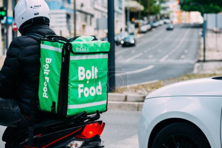 Foto de Lisboa, Portugal - 28 de abril de 2024: Bolt Food delivery rider, luciendo una brillante mochila verde aislada, se ve desde atrás mientras espera al lado de una carretera urbana concurrida - Imagen libre de derechos