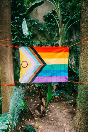 Una bandera del Orgullo LGBT, incluida la variante del Orgullo del Progreso, se exhibe en medio de una vegetación espesa y palmeras en un jardín tropical. La bandera está asegurada con cordones rojos
