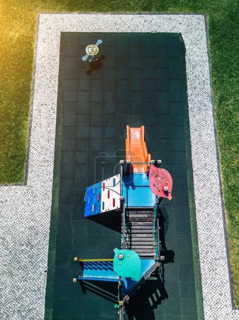 Luftaufnahme eines lebhaften Spielplatzes mit verschiedenen Freizeiteinrichtungen für Kinder, darunter eine Rutsche und Klettergeräte, an einem hellen, sonnigen Tag. Das Gebiet ist von ordentlich geschnittenem Gras und steinernen Wegen umgeben.