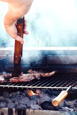 Eine Hand, die eine Zange benutzt, um ein Steak auf einem Grill zu drehen. Das Steak liegt auf einem Grillrost über heißen Kohlen, aus den Kohlen steigt Rauch auf. Die Hand trägt ein Armband.