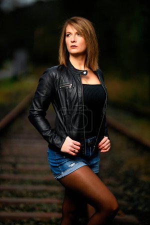 Photo for A young woman in a leather jacket poses on railway tracks, looking into the camera and the flash that illuminates the scene. View like Studiolight. Flash unleashed. - Royalty Free Image