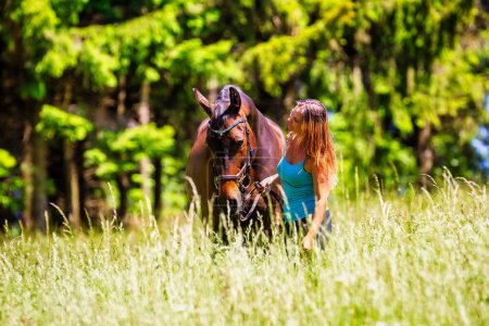 Una joven jinete con el pelo largo morena se para con su caballo en un alto prado de verano bajo el sol brillante.