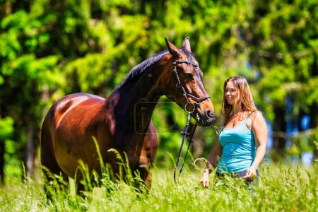 Photo for A young woman rider with long brunette hair stands with her horse on a high summer meadow in the bright sunshine. - Royalty Free Image