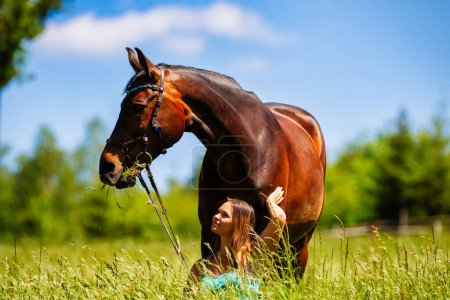 Photo for A young woman rider with long brunette hair stands with her horse on a high summer meadow in the bright sunshine. - Royalty Free Image