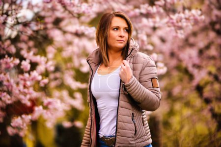 Photo for One young woman stands in a sea of cherry blossoms and smiles into the camera. Cherry blossoms on the tree in full pink bloom. - Royalty Free Image
