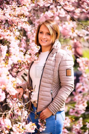Téléchargez les photos : Femme aux cheveux courts bruns avec des stries en jeans et gilet matelassé en portrait entre cerisiers en fleurs. - en image libre de droit