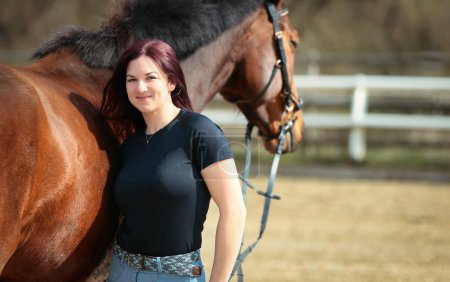 Photo for Woman in civilian clothes stands with her horse on the riding arena. She smiles into the camera and strokes her horse. Upper body portraits. - Royalty Free Image