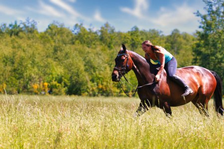 The picture shows a young woman on her horse in a summer meadow on a sunny summer day. ridden without a saddle with only a bridle, in the background forest and blue sky.