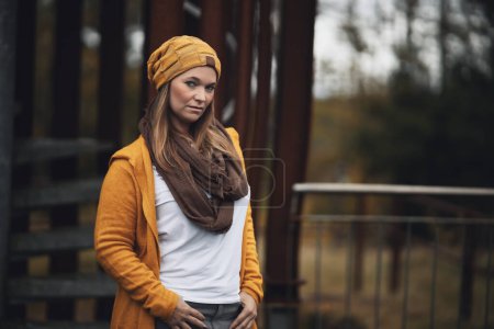 Photo for Woman brunette in portraits. She is in her mid-thirties and is dressed in a jeans, yellow knitted coat, t-shirt and a yellow hat. She stands in front of a metal scaffolding in nature. Landscape format with space for text. - Royalty Free Image