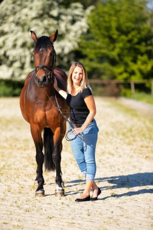 Photo for A young woman stands with her horse on a sunlit riding arena. In the background, blooming trees and green nature, model isolated in front of blur, brown horse and woman dressed with a black shirt and jeans. - Royalty Free Image