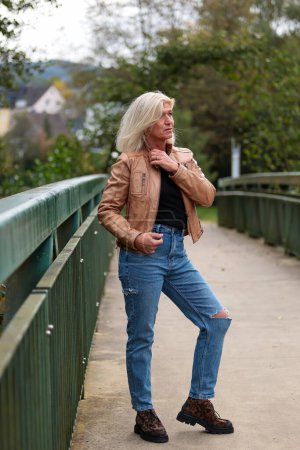 Foto de Una mujer mayor rubia de unos 50 años vestida con una chaqueta de cuero marrón, jeans y una camisa negra posando exuberantemente sobre un puente peatonal en un parque. - Imagen libre de derechos