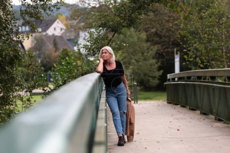 Foto de Una mujer mayor rubia de unos 50 años vestida con una chaqueta de cuero marrón, jeans y una camisa negra posando exuberantemente sobre un puente peatonal en un parque. - Imagen libre de derechos