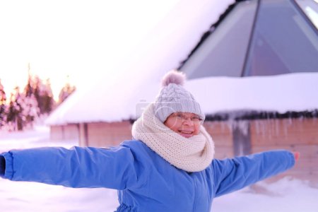 Foto de Mujer divertida en puestos de ropa de invierno cerca de casa de madera con techo de cristal, moderno iglú cubierto de nieve finlandesa en el norte de Laponia, pueblo de invierno, tradicionalmente turismo con polo norte ártico finlandés - Imagen libre de derechos