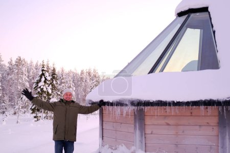 Foto de Gracioso hombre maduro en ropa de invierno se encuentra cerca de casa de madera con techo de cristal, moderno iglú cubierto de nieve finlandesa en el norte de Laponia, pueblo de invierno, tradicionalmente turismo con polo norte ártico finlandés - Imagen libre de derechos