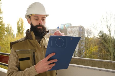 professional male foreman in helmet during inspection in construction site, bearded man in uniform, contractor, architect on building site writing on clipboard, inspect project performance