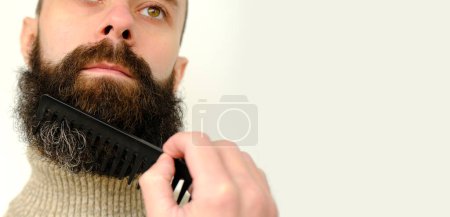Foto de Primer plano de la cara masculina, joven barbudo y hombre bigotudo, chico con 25 - 30 años peina su barba gruesa, concepto de bigote y barba productos de cuidado de rastrojos, higiene de la línea del cabello en la parte inferior de la cara - Imagen libre de derechos
