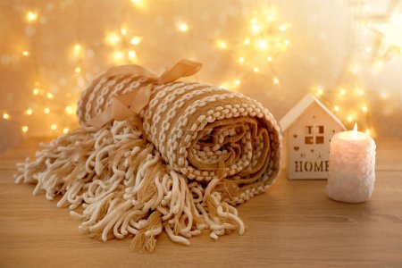 beautiful cozy creamy plaid with fringe lies rolled up on light table against festive background with bokeh of garlands, wooden house with candle, concept of sweet house, warm cute gift for holiday