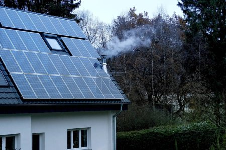 Foto de Parte de acogedora casa blanca en invierno en pueblo europeo, paneles solares instalados en el techo, el humo proviene de la chimenea, calefacción económica ecológica autónoma de la vivienda, ecología verde - Imagen libre de derechos