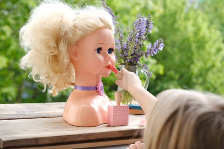 Photo for Girl's hands imitates painting lips doll, playing with set of stylist, beautician, hairdresser on wooden table in garden, on balcony, simulation games in profession, makeup artist - Royalty Free Image