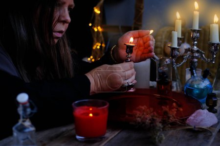 female witch holds candle in candlestick in her hands, conjures in dark room, magic items on table, concept of Halloween party, alternative medicine, esoteric practices, seance in salon of medium
