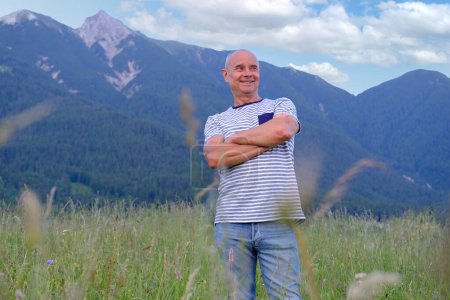 Photo for Mature man, senior 60 years old standing in mountains above city, smiles, Alps are behind him, green grass in meadow, concept of enjoy life in old age, travel, active lifestyle, human happiness - Royalty Free Image