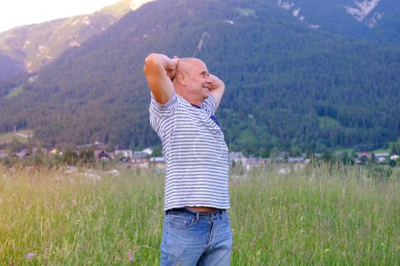 Photo for Mature man, senior 60 years old standing in mountains above city, smiles, Alps are behind him, green grass in meadow, concept of enjoy life in old age, travel, active lifestyle, human happiness - Royalty Free Image