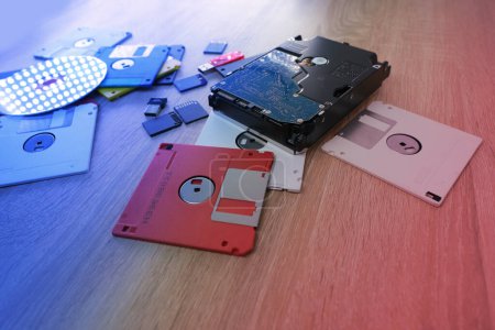 Foto de Dispositivos de almacenamiento de datos electrónicos retro vintage de los años 80, 90, disco cd, unidades flash, disco duro moderno dispersos en la mesa. Pila de disquetes en gris, negro, azul, amarillo, rojo, blanco, luz de fondo roja - Imagen libre de derechos