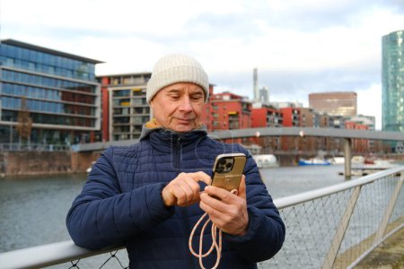 Photo for Positive charming mature man in European City Frankfurt using modern phone with smile, highlighting role of gadgets in everyday business communication, Mobile Usage, Digital Lifestyle - Royalty Free Image