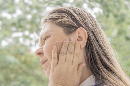 Foto de Mujer caucásica madura 50 años cogida de la mano para dolor de oído, primer plano de la cara femenina con dolor de expresión facial, zumbido en los oídos, acúfenos, inflamación del oído medio, trauma acústico - Imagen libre de derechos