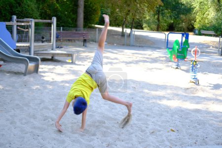 child, boy 7-8 years old in yellow T-shirt, shorts performs barefoot combination of wheel in side somersault, Salto on playground in summer park, concept motor activity of children, active lifestyle