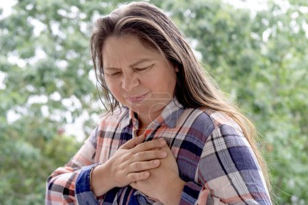 Frau im Alter von 40-45 Jahren in kariertem Hemd hält Herz, plötzliche Brustschmerzen, aus nächster Nähe weibliches Gesicht mit Gesichtsausdruck leiden, ischämische Herzkrankheit, arterielle Hypertonie, Myokarditis oder Arrhythmie