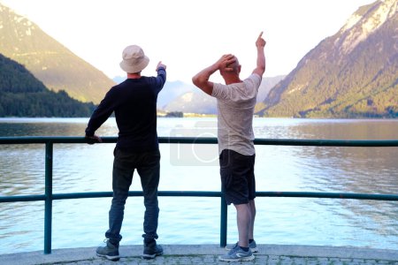 zwei Männer stehen auf Aussichtsplattform, bewundern Berglandschaft, Achensee in Österreich, grüne Berge erheben sich über ruhiges Weitwasser, Urlaub am Stausee, gemeinsame Reise, Erholungsort Tirol