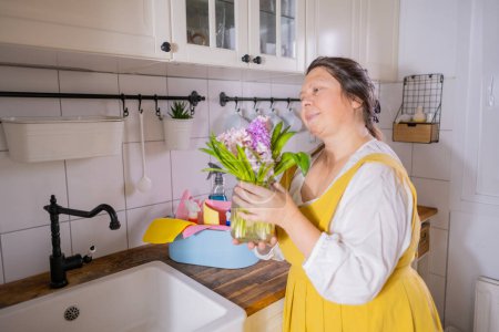 reife Frau mit Frühlingsstrauß aus weißen und lila Hyazinthen in den Händen, während sie in der Küche gelbe Schürze trägt, Frühjahrsputz zu Hause, Zufriedenheit in Frische und Schönheit Frühling