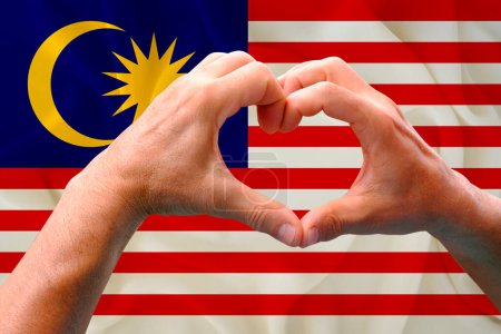 close-up of male hands in heart form against background of silk national flag of Malaysia, patriots of country concept, Independence day, travel, international cooperation