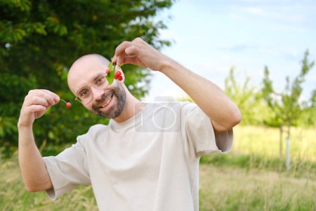 closeup male hands holds ripe red cherries, happy young charismatic man in beige t-shirt smiles cheerfully, concept vegetarian food, wholesome healthy eating, food enriched with vitamins