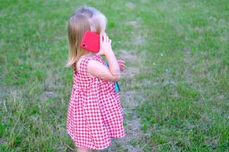 Photo for Small child, 2-year-old girl with white hair in red dress holds smartphone near her ear, communicates with friends, parents, concept of childhood, previously ability to handle modern technologies - Royalty Free Image