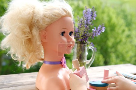 girl's hands are applying makeup on doll, playing with set of stylist, beautician, hairdresser on wooden table in garden, on balcony, simulation games in profession, makeup artist
