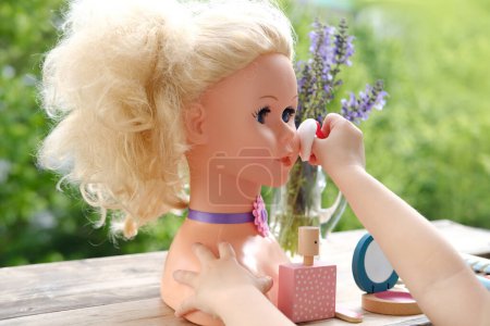 girl's hands are applying makeup on doll, playing with set of stylist, beautician, hairdresser on wooden table in garden, on balcony, simulation games in profession, makeup artist
