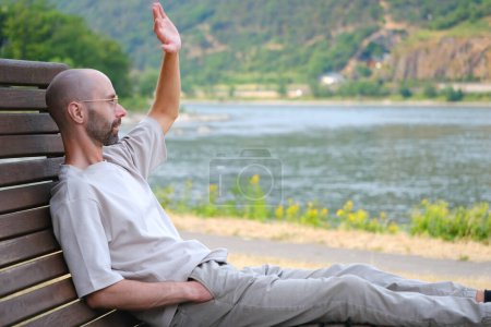 young man 30 years old sites comfortably on wooden park bench on banks of river, lake, resting on shore in harmony with nature, raised hand in greeting, nature therapy, enjoying nature