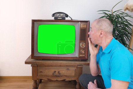 älterer Mann in blauem Poloshirt sitzt auf dem Boden vor dem alten analogen Retro-Fernseher, Fernseher, fasziniert von aufregendem Programm, Green Screen Attrappe, Ausdruck der Überraschung, Älterer Lebensstil