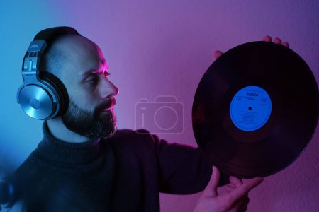 close up of Young man with beard in headphones holds vinyl disc in his hands, contemplatively looking in neon purple and green light, DJ culture, Neon-lit contemplation, crimson bright backlighting