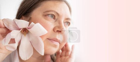 aging woman with magnolia flower looks herself in mirror, noticing wrinkles, changes in facial contour and sagging eyelids, cosmetic anti-aging procedures, natural process growing older